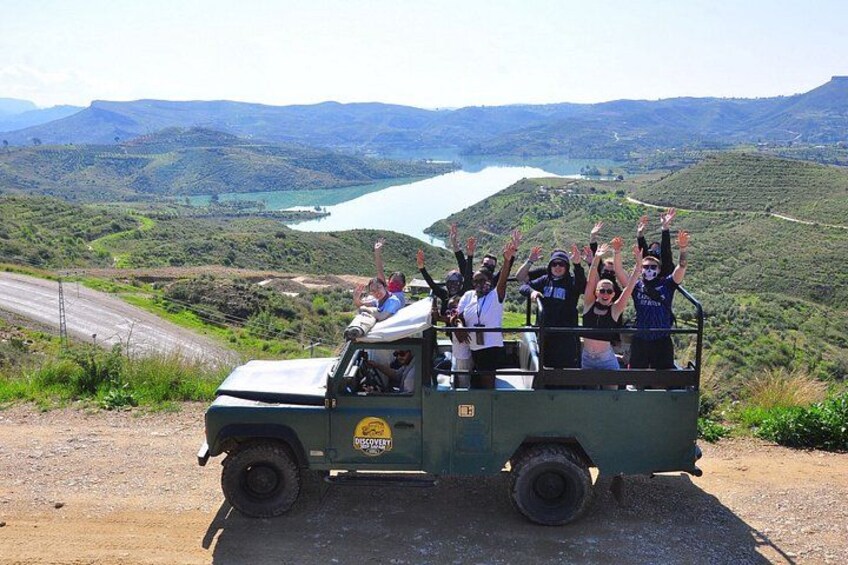 A green jeep with passengers raising their hands, overlooking a lake and hilly landscape. 1