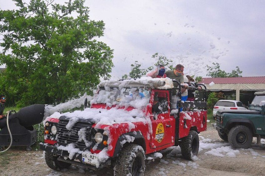 A red off-road vehicle covered in foam, parked on a gravel path with trees in the background. 5