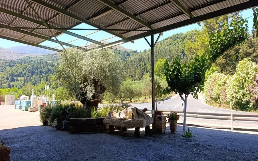 A covered outdoor area with a gravel ground, a stone bench, potted plants, and a view of a road and forested hills. 4
