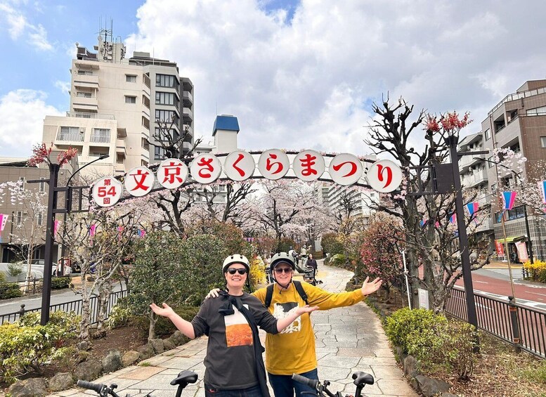 Two people on a bike path with cherry blossoms and a sign with Japanese characters. 5