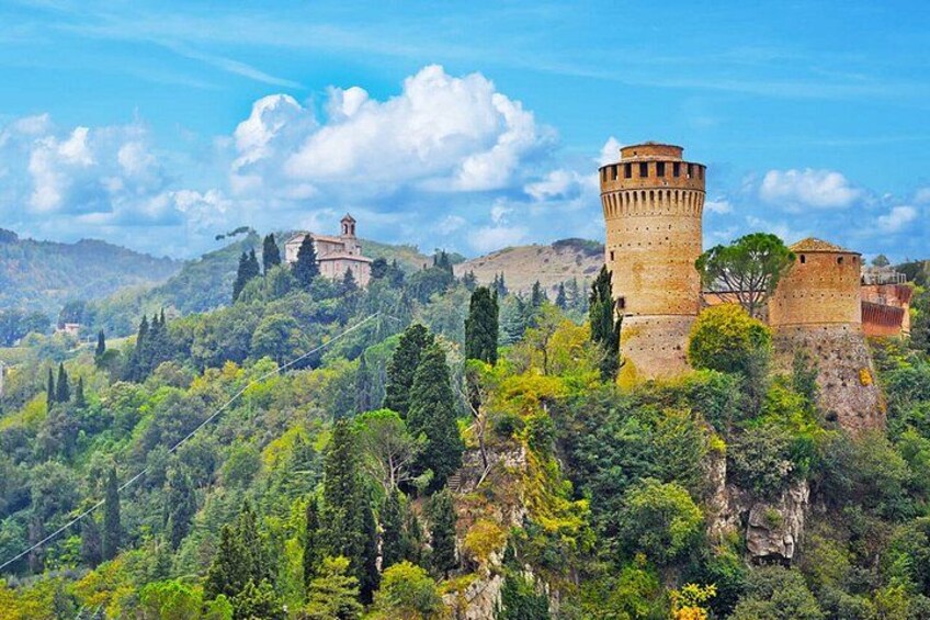 A historic tower and fortress on a hill, surrounded by lush greenery and other buildings. 1