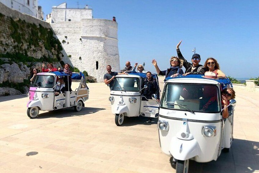 Three small vehicles with passengers, parked in front of a white building with a wall and a flag. 3