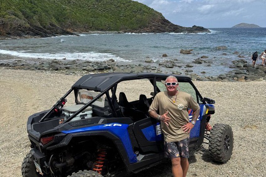 Un homme se tient à côté d’un véhicule tout-terrain bleu sur une plage rocheuse, avec l’océan et des collines en arrière-plan. 5