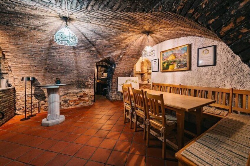 A subterranean dining area with wooden benches and a stone table, featuring a thatched ceiling and rustic brick walls. 5