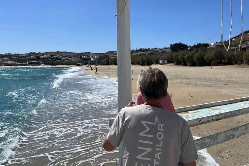 En person i en skräddarmässig uniform står vid en strand, med vågor som slår in mot stranden och en klar himmel ovanför. 5