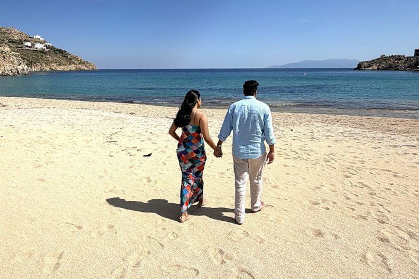 Un couple se promène sur une plage de sable, avec une eau calme et des formations rocheuses en arrière-plan. 1