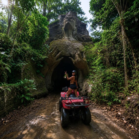 A person on a red ATV entering a cave-like structure surrounded by dense forest. 3