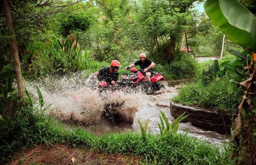 Two people on a red ATV navigating through a shallow water crossing surrounded by dense greenery. 1