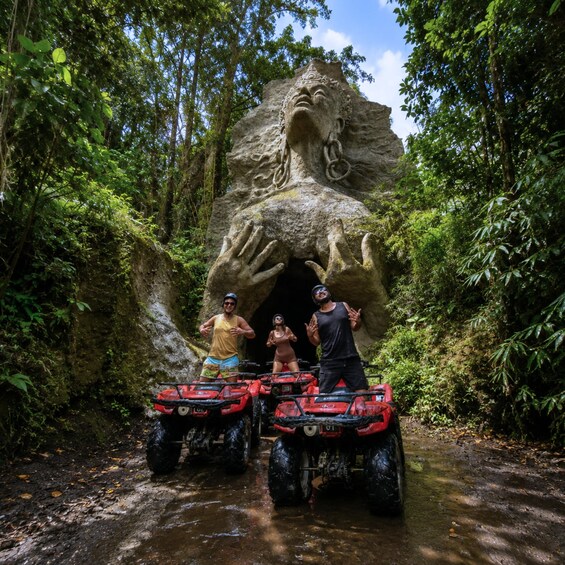 Three people on ATVs in front of a large rock formation with a cave-like entrance. 4