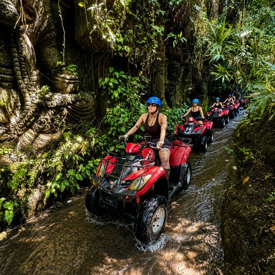 A group of people on red all-terrain vehicles navigating a narrow, water-filled path through a dense, tropical forest. 1
