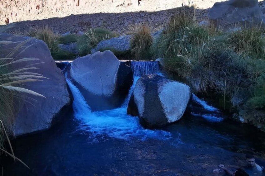 A small waterfall with large rocks, surrounded by grass and desert landscape. 1