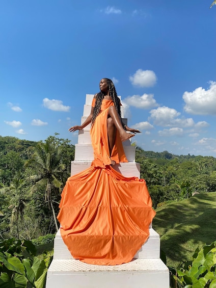 A person in an orange dress sitting on a white structure with a lush green landscape in the background. 2