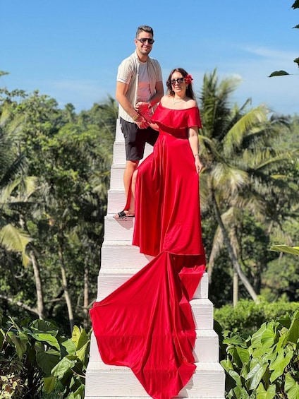 A couple standing on a white staircase with a red dress and a white shirt, surrounded by greenery and palm trees. 3