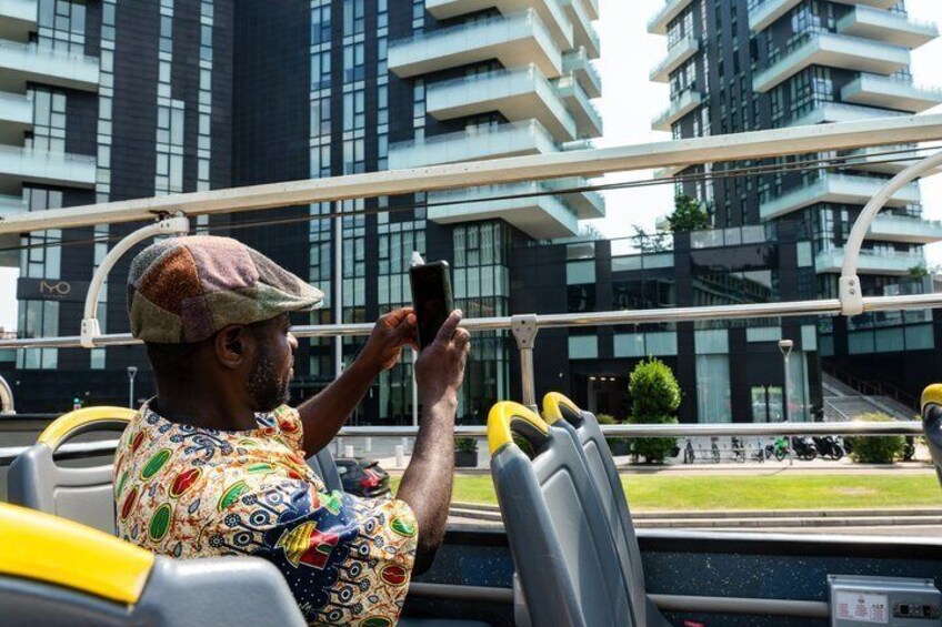 Una persona tomando una foto con un teléfono inteligente, sentada en un vehículo con asientos amarillos y grises, con edificios modernos al fondo. 4