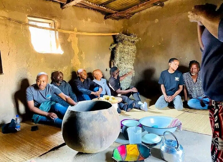 A group of people sitting in a rustic room with a large pot, bowls, and a water container on the floor. 3