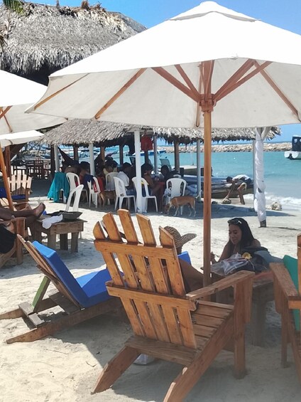 A beach with wooden chairs and umbrellas, people sitting and relaxing, and a thatched-roof structure in the background. 2