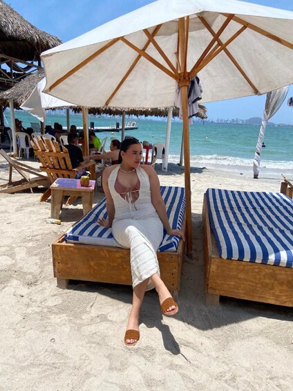 A woman sitting on a wooden lounge chair under a large umbrella on a sandy beach with a city skyline in the background. 5