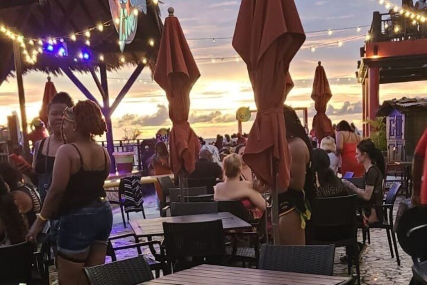 Un bar en plein air très fréquenté, avec des tables et des parasols, offrant une vue sur le coucher du soleil. 3