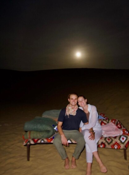 Two people sitting on a bed in a desert at night with a full moon in the background. 2
