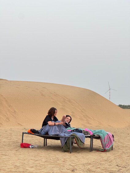 Two people resting on a metal bench in a desert with sand dunes and a wind turbine in the background. 5