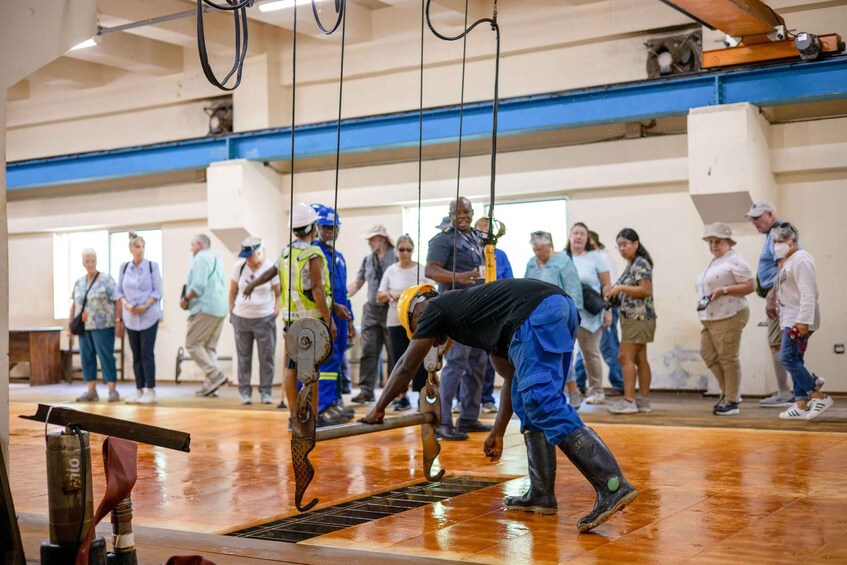 A person in blue overalls working on a mechanical device in a room with a wet floor, surrounded by onlookers. 2