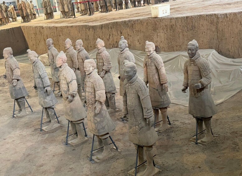 Terracotta soldiers lined up in rows, some standing and some kneeling, with a large earthen mound in the background. 2