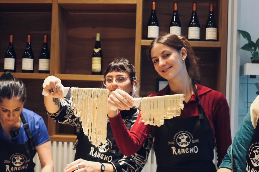 Three individuals wearing aprons with 'Rancho' branding, preparing food in a kitchen with wine bottles in the background. 1
