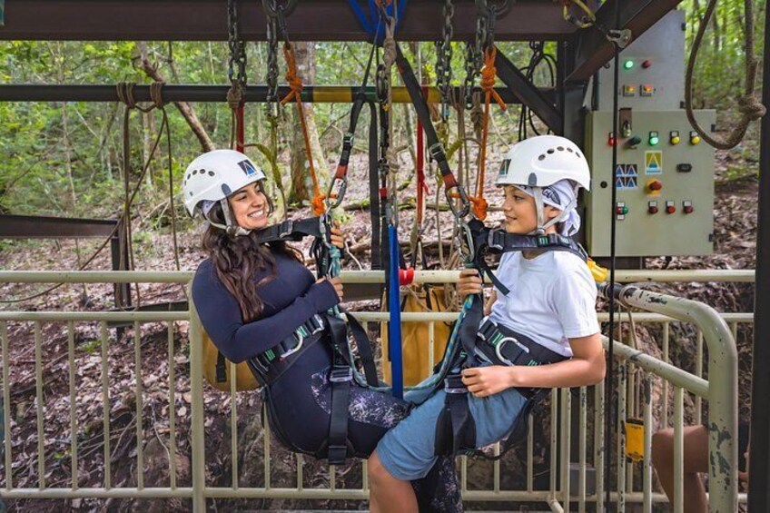 To personer på en zipline, iført hjelme og seler, med en skov i baggrunden. 3