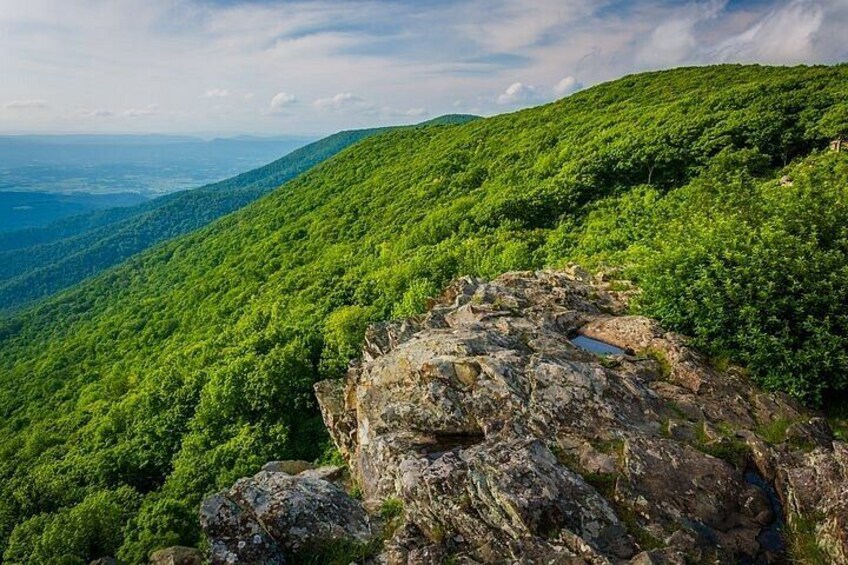 A mountainous landscape with dense green forest, rocky outcrops, and a small water body. 4