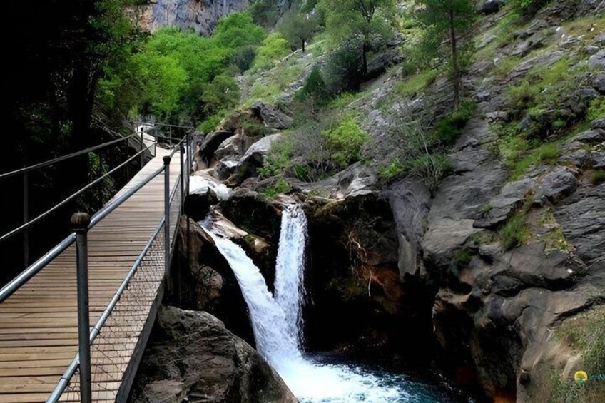 A wooden bridge over a waterfall with surrounding greenery and rocky terrain. 2