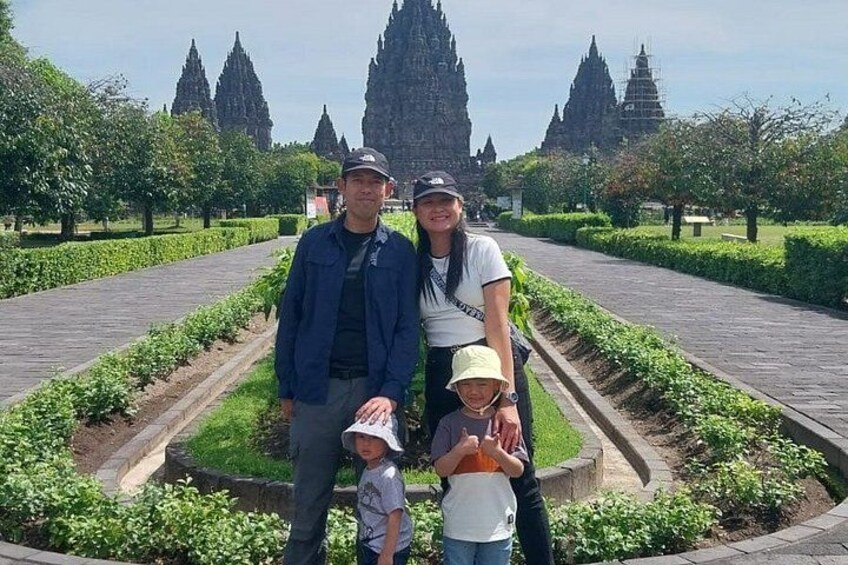 A family of four standing in a landscaped garden with a pathway, surrounded by greenery and a large temple structure in the background. 4