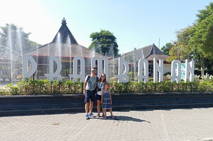 A family standing in front of a large sign with the text 'DRAMBANAN' and a water fountain in the background. 3