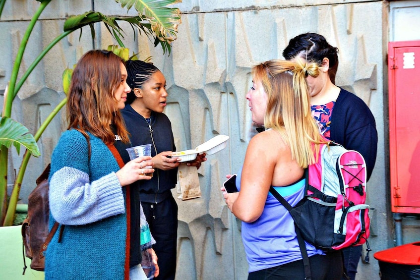 A group of people standing outdoors, one holding a paper plate and a drink, another with a smartphone, and a third with a pink backpack. 2