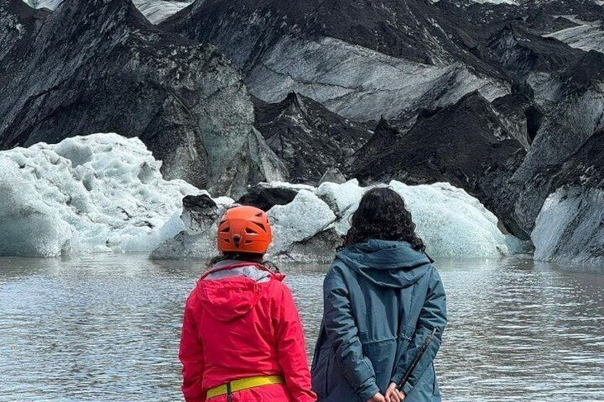 Two people standing by a glacier with ice formations and rocky terrain in the background. 1