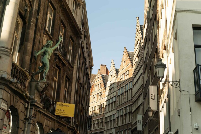 A narrow street with historic buildings, a statue, and a sign for De Melleer. 3
