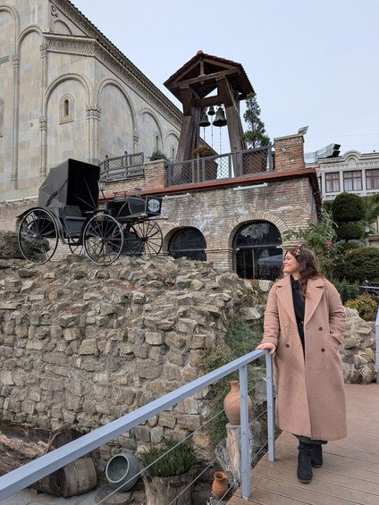 A person standing on a stone wall with a wooden bell tower and a horse-drawn carriage in the background. 3