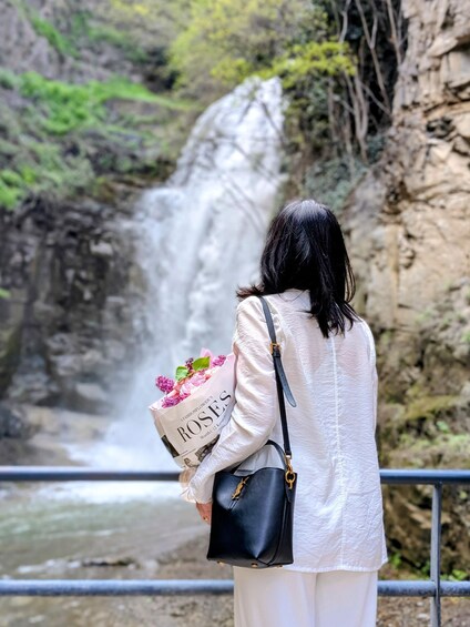 A person holding a bouquet of flowers and a bag, standing by a waterfall. 1