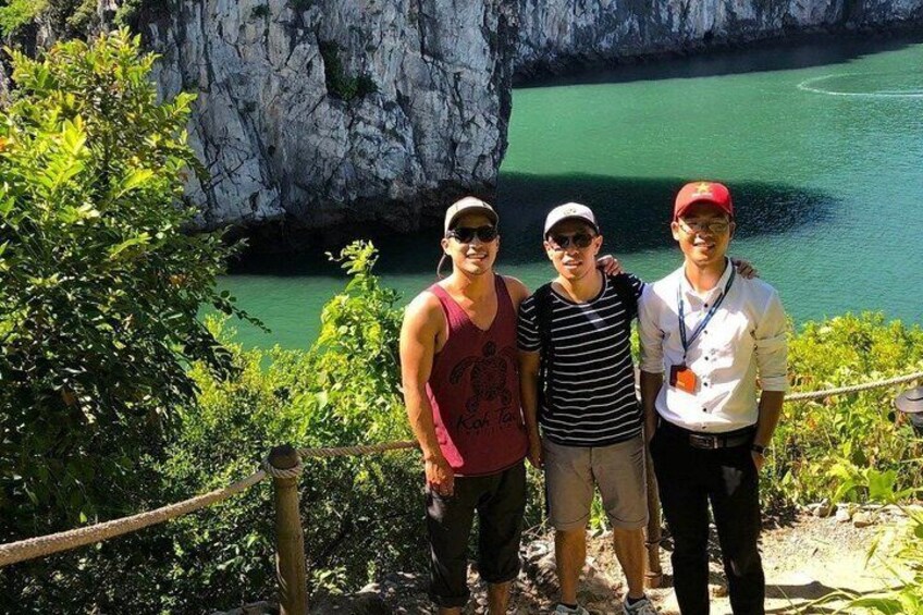 Three people standing on a trail with a rocky cliff and a green body of water in the background. 4
