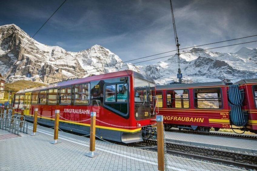 A red Jungfraubahn train at a station with snow-covered mountains in the background. 1