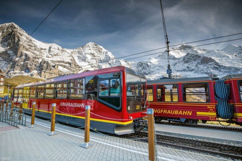 A red and yellow train at a station with snow-capped mountains in the background. 1