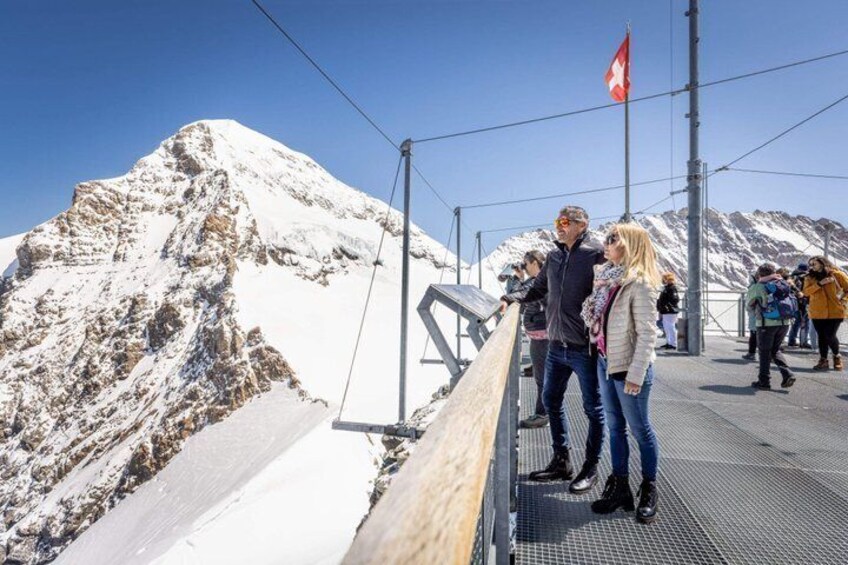 A snowy mountain with a Swiss flag, a metal railing, and people on a viewing platform. 3