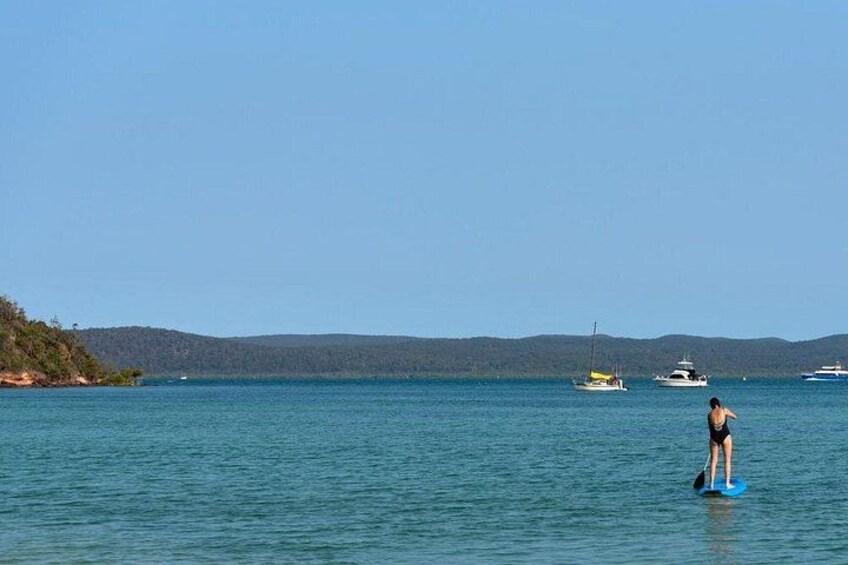 A person surfing on a calm sea with boats in the background and a hilly landscape. 2