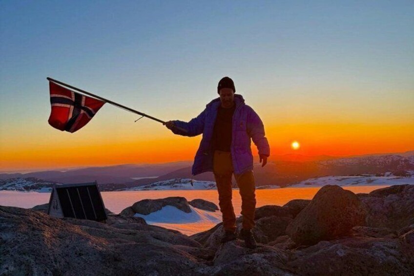 A person standing on a rocky terrain with a flag, a sunset in the background, and snow-covered mountains. 4