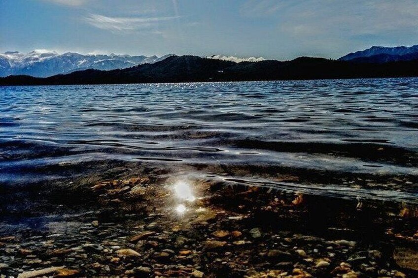 A lake with clear water reflecting sunlight, pebbles on the shore, and mountains in the background. 3