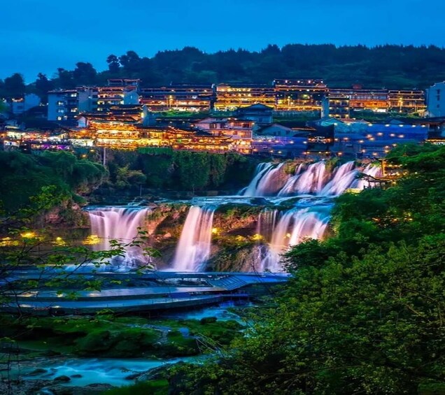 A waterfall cascading into a river surrounded by lush greenery and traditional buildings at dusk. 1