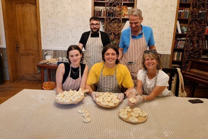 Four people wearing aprons, working with dough on a table in a room with wooden furniture and bookshelves. 1