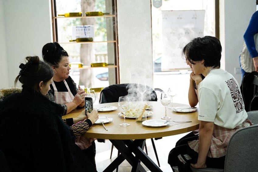 A group of people sitting around a table with food and drinks, in a room with a window and shelves with bottles. 5