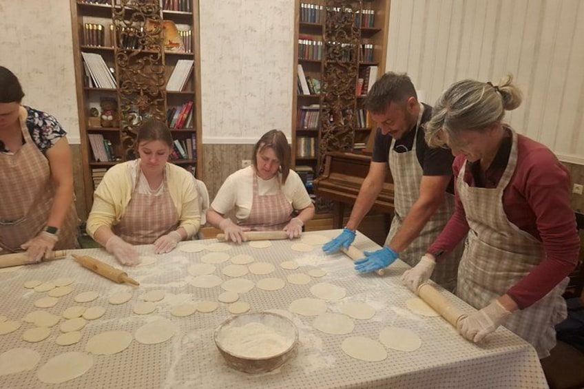 A group of people rolling out dough on a table in a room with bookshelves. 2
