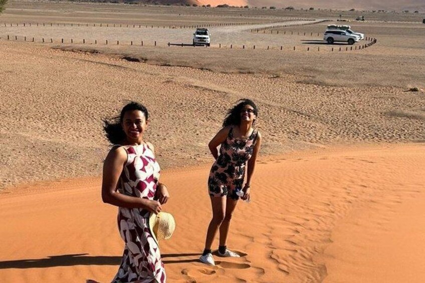 Dos personas de pie sobre una duna arenosa con un paisaje desértico y vehículos al fondo. 3