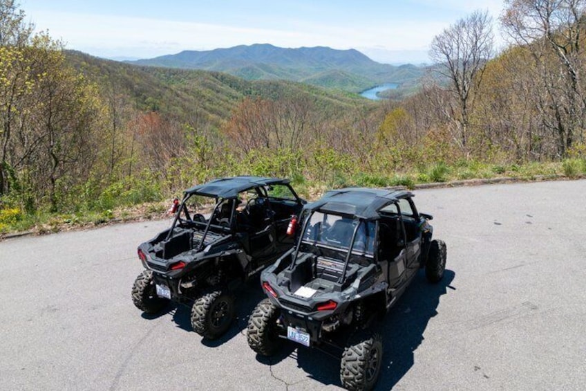 Two off-road vehicles parked on a paved road with a scenic view of a river and mountains in the background. 3